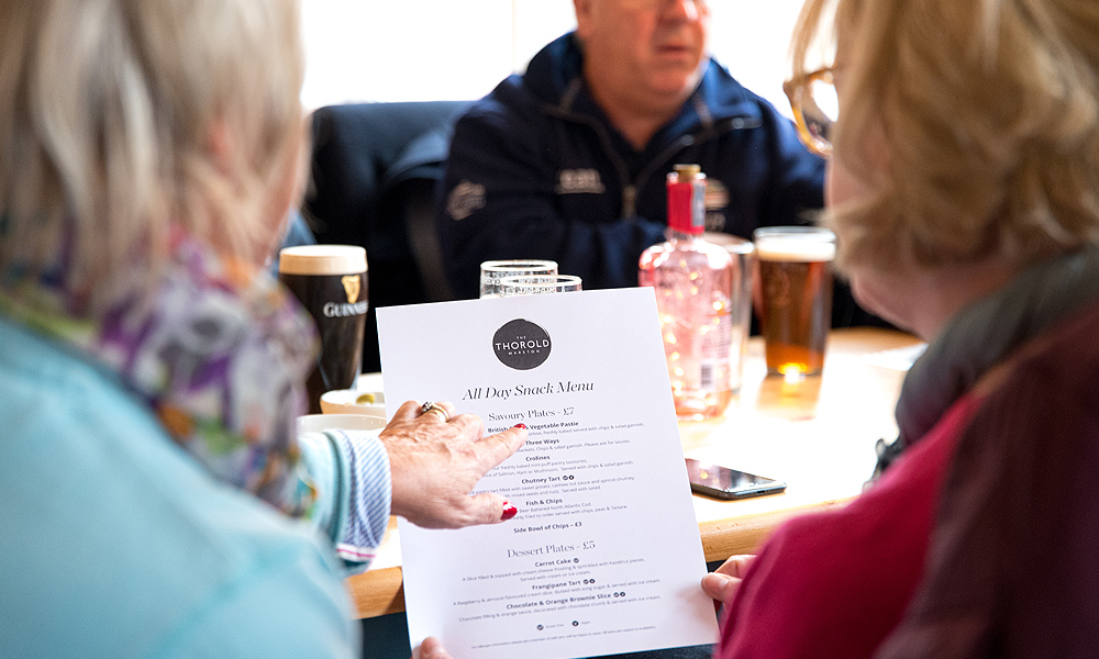 This image shows two people seated at a table in what appears to be a café, with one person pointing at an "All Day Snack Menu." In the background, you can see drinks on the table and other customers.