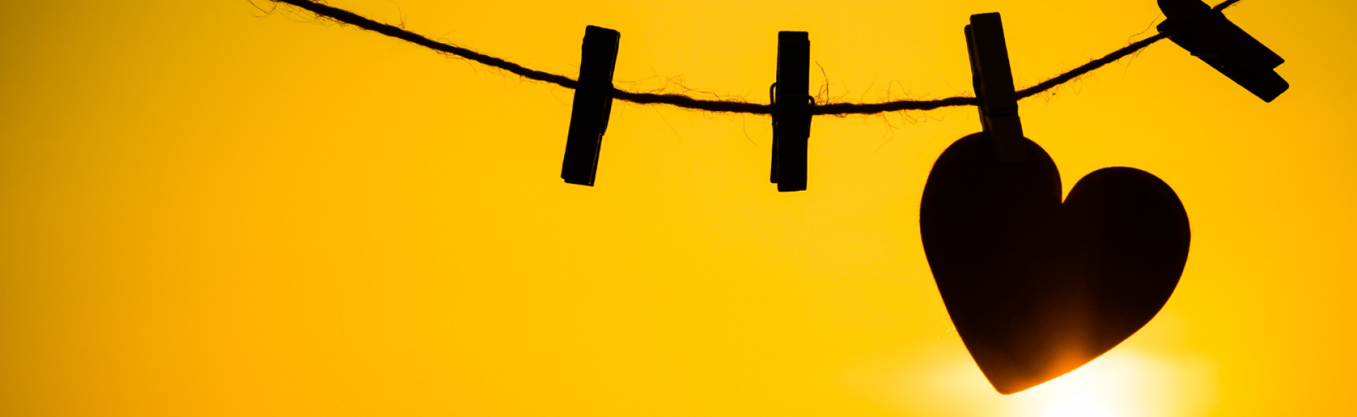 Silhouette of a heart shape being held on a string by a clothing peg. The background shows the sun shining against a yellow sky