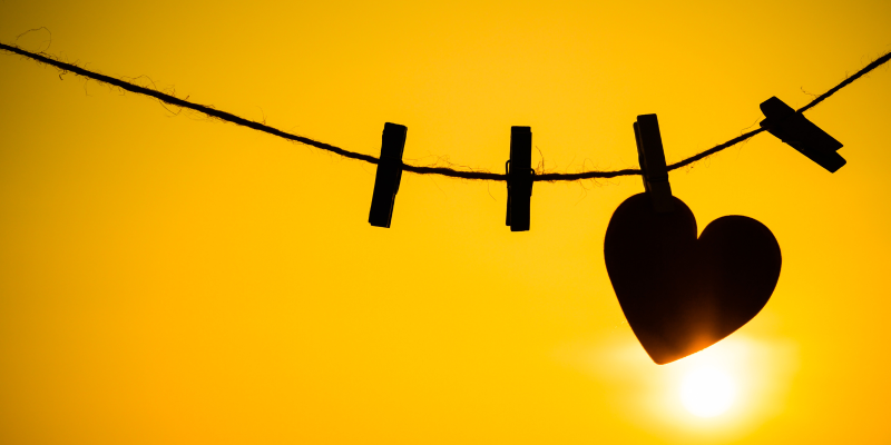 Silhouette of a heart shape being held on a string by a clothing peg. The background shows the sun shining against a yellow sky