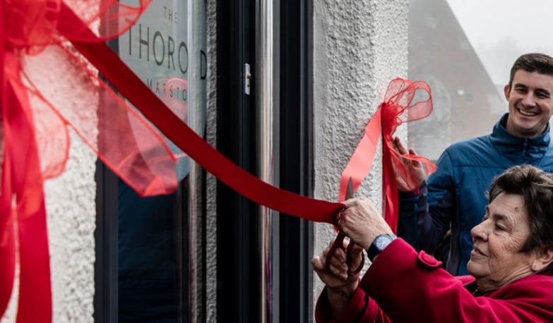 This image shows an older woman cutting a bright red ribbon in front of a building, symbolizing the official opening of a new space. She is smiling as she does so, with another person, possibly a younger man, smiling in the background and observing the event. 