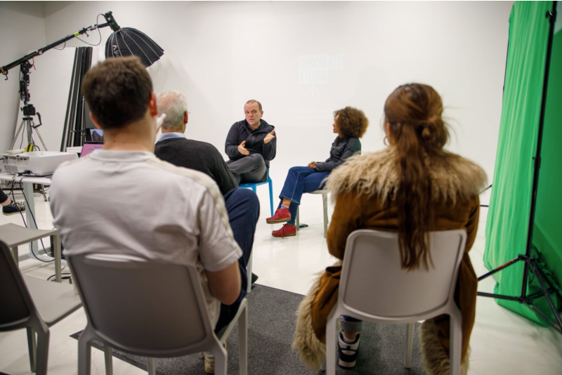 A small group of people sits in a studio, attentively listening to two individuals engaged in a discussion. The setting includes professional lighting equipment, a green screen, and a projector, indicating a media or film-related event. The "Chocolate Films" logo is faintly visible on the white backdrop.